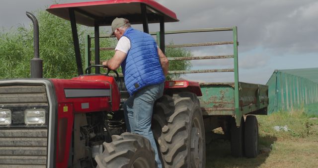 Senior farmer wearing a blue vest climbing into a red tractor on a rural farm with green trailer and cloudy sky. Perfect for illustrating agricultural work, rural life, senior lifestyle, and farm equipment usage.