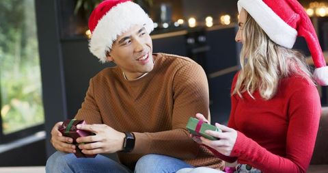 Diverse couple exchanging christmas gifts on couch wearing santa hats and cozy sweaters