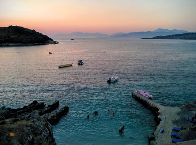 Boats float on a tranquil ocean as the sun sets, creating a peaceful scene. People are in the water near the shore, enjoying the evening. Ideal for travel and leisure websites, blogs about nature, and promotions of beach vacations.