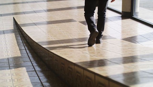Walking man crossing raised patterned tile platform in sunlit glass lobby with window shadows