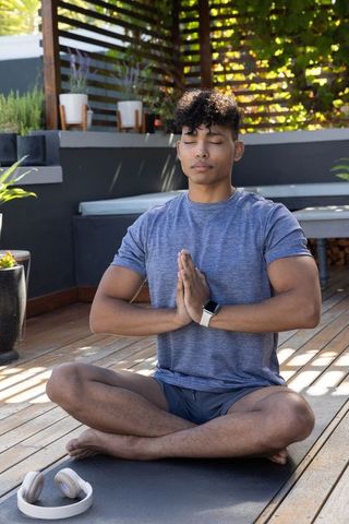 Young man meditating with hands in prayer on outdoor yoga mat with smartwatch and headphones