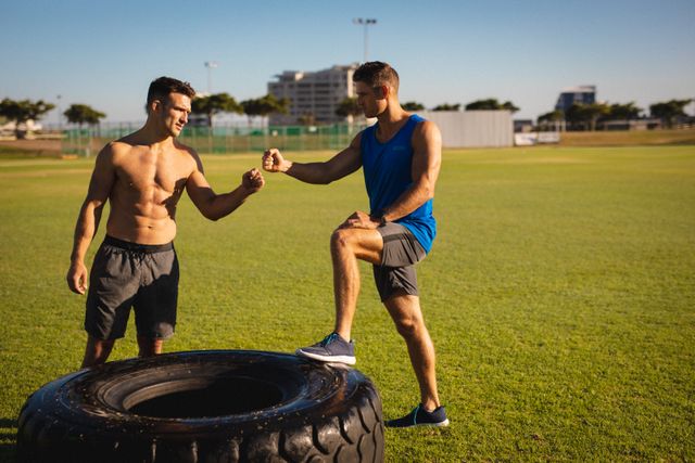 Two fit caucasian men standing at tire, fist bumping outdoors. cross training for fitness at a sports field.