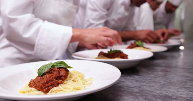 Group of professional chefs wearing white uniforms plating spaghetti dishes with meat sauce and garnish in a professional kitchen. Ideal for content related to culinary arts, cooking classes, restaurant advertisements, chef training, food industry promotions, and gourmet dining experiences.