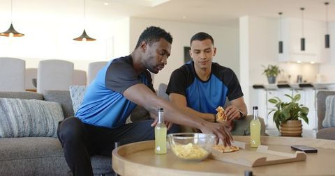 Diverse friends relaxing on sofa sharing pizza and drinks while watching game at home