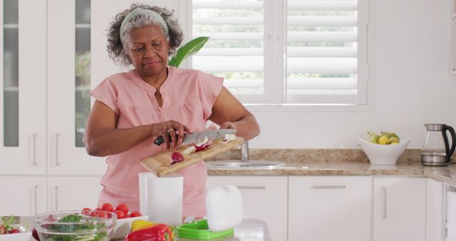 Senior African American woman cutting vegetables in a bright, modern kitchen. She appears relaxed and focused, showcasing healthy eating habits and home cooking. Great for use in advertisements for kitchen products, healthy lifestyle promotions, senior living articles, or representation of everyday life for older adults.