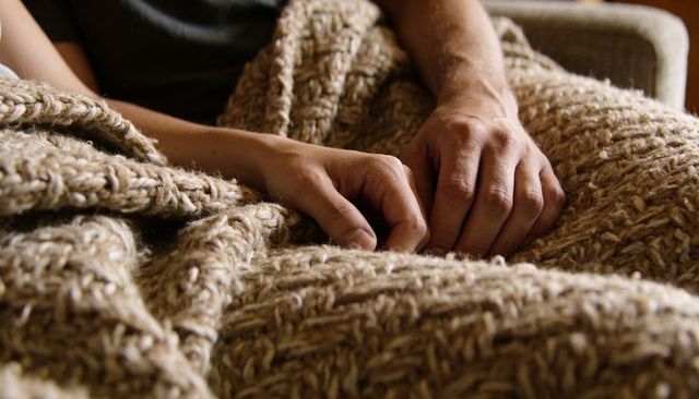 Resting hands under chunky beige knit blanket on sofa closeup for cozy comfort and warmth