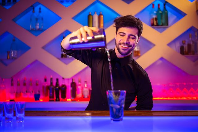 Confident bartender pouring cocktail into glass at modern bar counter with illuminated background. Ideal for use in articles about nightlife, bar culture, mixology, and professional bartending. Suitable for promoting bars, nightclubs, and cocktail recipes.