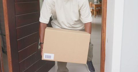 African man carrying cardboard delivery box through home doorway into interior
