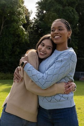 Diverse female partners embracing in park wearing knit sweaters and jeans with smartwatch