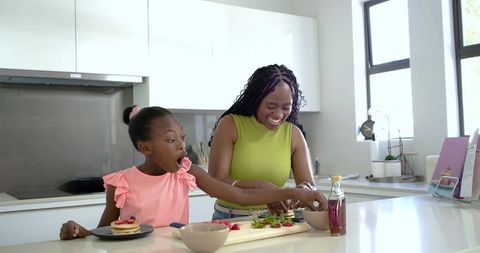 Black mother and daughter preparing pancakes and arranging strawberries on sunlit kitchen island