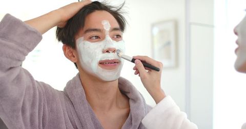 Partner applying facial mask to smiling man in gray robe at bathroom vanity