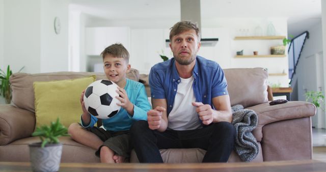Father and son are seen sitting on a sofa in the living room, engrossed in a soccer game on the TV. The boy is holding a soccer ball, and both are displaying expressions of excitement. This image can be used to illustrate family bonding moments, parental involvement in children's interests, and fan enthusiasm for sports. Ideal for family lifestyle, parenting, sports, and home entertainment themes.