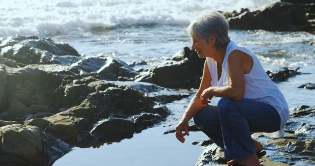 Senior woman enjoying a leisurely day on a rocky beach, wearing casual jeans and a sleeveless top. She appears tranquil as she looks into a tide pool, with ocean waves crashing gently in the background. Ideal for depicting themes of tranquility, connection with nature, and relaxation. Great for use in travel, retirement, and lifestyle promotions or articles.