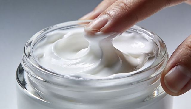 Pressing fingertip into glass jar of white moisturizer showing creamy texture and shine
