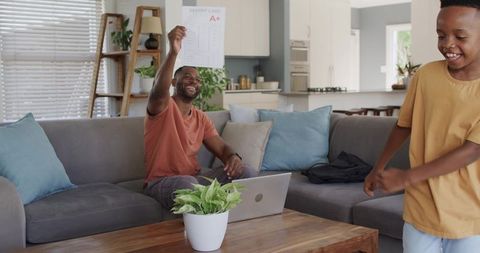 African American father celebrating son holding A+ report card on living room sofa
