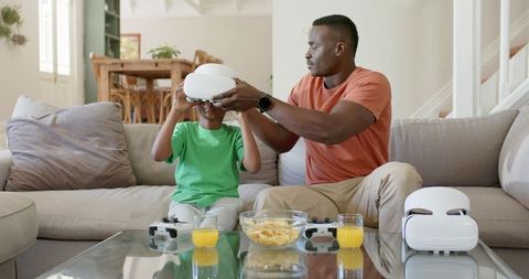 Father adjusting VR headset on son on couch, game controllers and snacks, family bonding