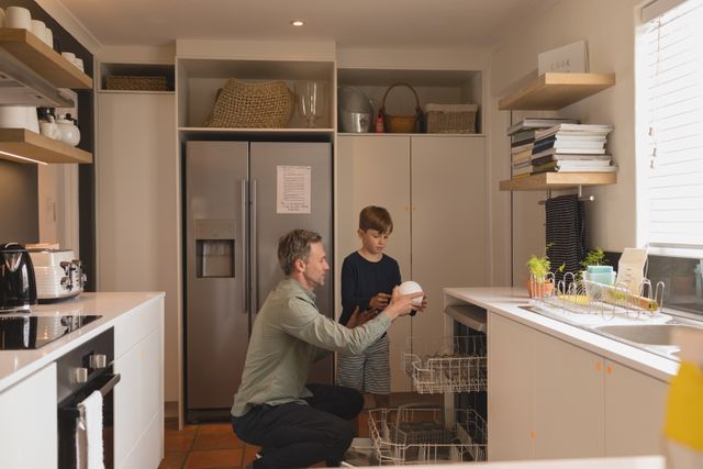 Father and son working together to unload the dishwasher in a modern kitchen. This image can be used to depict family bonding, teamwork in household chores, and daily domestic life. Ideal for articles or advertisements related to family life, home appliances, and parenting.