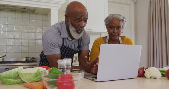 Senior couple looking at a laptop together while cooking in their kitchen. Various fresh vegetables like cabbage, bell pepper, and carrots are on the counter. They may be following an online recipe or attending an online cooking class. Suitable for content related to healthy eating, senior citizens using technology, online learning, or home cooking.