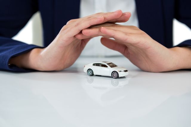 Businesswoman's hands forming a protective gesture over a toy car, symbolizing insurance and safety. Ideal for use in articles, advertisements, and presentations related to car insurance, vehicle protection, risk management, and business security concepts.