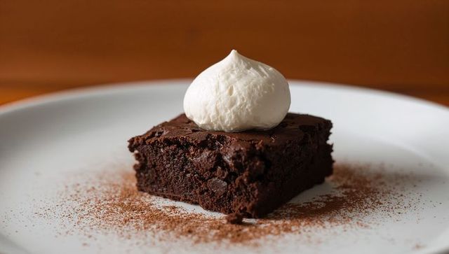 Fudgy chocolate brownie sitting on white plate showing whipped cream dollop and cocoa dust