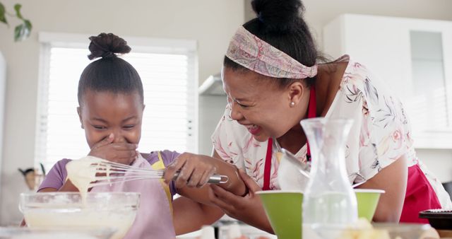 Mother and daughter are laughing and bonding while baking in a kitchen. Mother holds a whisk and the daughter has a playful smile. They are enjoying quality time together, creating a joyful atmosphere. Ideal for concepts related to family bonding, parent-child activities, cooking at home, and happy family moments.