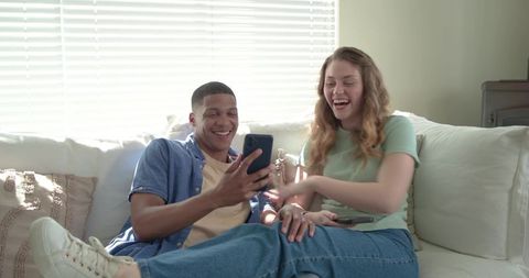 Young couple laughing while scrolling smartphone on bright living room sofa
