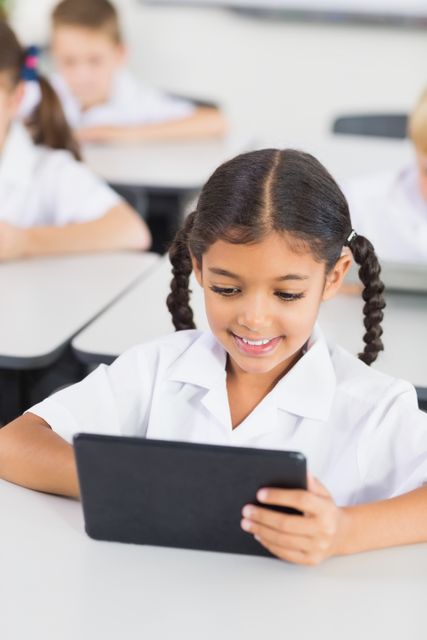 Young schoolgirl using a digital tablet in a classroom setting, smiling while focusing on the screen. Other students can be seen in the background, also engaged in their tasks. This image can be used for educational websites, articles about technology in education, school advertisements, or resources for teachers and parents to showcase the integration of digital tools in modern learning environments.