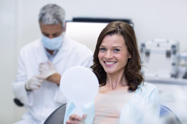 Patient is smiling while checking her teeth in a mirror at a dental clinic. Dentist in background preparing for treatment. Ideal for use in healthcare, dental care, and medical service promotions.