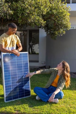 Diverse homeowners inspecting large solar panel on suburban lawn planning residential renewable ener