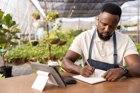 African american greenhouse entrepreneur writing notes at desk with tablet and plants