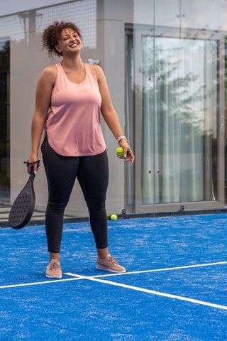 Smiling african american woman playing padel on bright blue court in pink activewear
