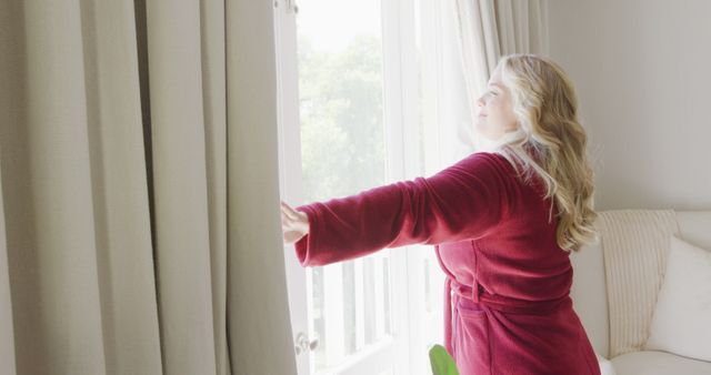 Happy plus size caucasian woman in bathrobe drawing bedroom curtains on sunny morning. Lifestyle, morning, free time, wellbeing and domestic life, unaltered.