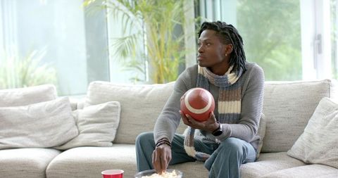African American man sitting on sofa holding football and reaching for popcorn