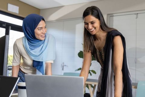 Collaborating women leaning over laptop in modern office, woman wearing hijab and tunic