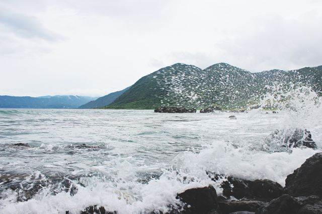 Waves crashing on a rocky shoreline with mountains in the background. This image highlights the power and beauty of nature, making it suitable for travel brochures, adventure blogs, and ecological campaigns.