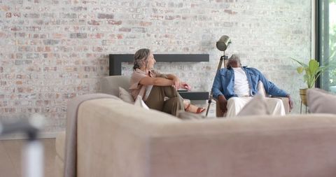 Relaxed multiracial couple chatting in sunlit minimalist living room with exposed brick wall