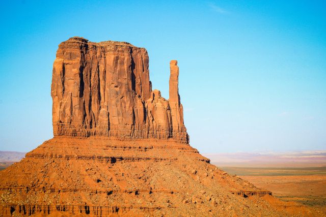 The image depicts a prominent rock formation in a vast desert landscape, illuminated by bright daylight. This striking scene, reminiscent of Monument Valley, showcases red sandstone formations against a clear blue sky. Ideal for travel publications, nature-themed websites, and geological studies, it captures the essence of the American Southwest’s natural beauty.