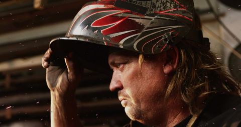 Mature welder lifting helmet amid glowing sparks in gritty metal fabrication workshop