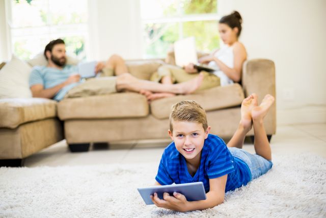 Young boy lying on carpet using digital tablet while parents sit on sofa with their own devices in background. Ideal for illustrating modern family life, technology use, and family bonding moments.