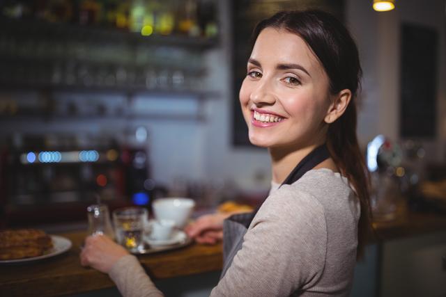 Young female waitress standing at a coffee shop counter, wearing an apron and smiling warmly. Ideal for use in advertising hospitality or food service industries, promoting careers in customer service, or illustrating friendly service in cafes and restaurants.