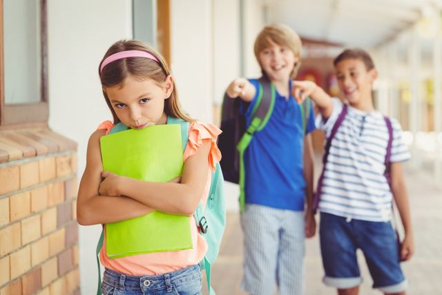 Sad schoolgirl holding folder while two classmates tease her in school corridor. Useful for illustrating issues like bullying, peer pressure, and emotional well-being in educational settings. Highlights the impact of bullying and the need for supportive school environments.