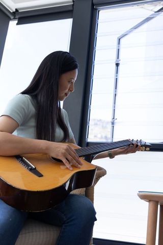 Sunlit acoustic guitar practice by mature african woman sitting in woven armchair, focused