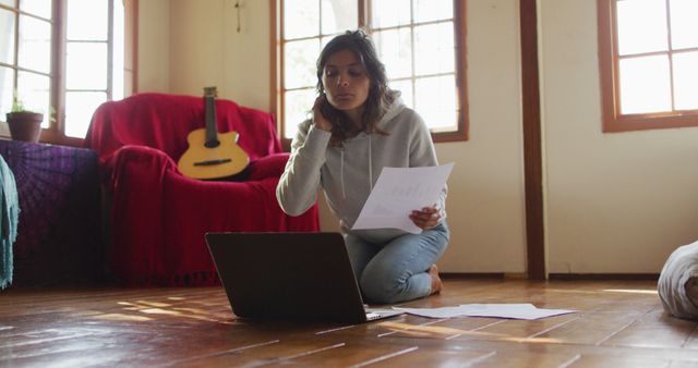 Young woman in casual clothing focusing on her laptop, reviewing documents. She is seated on the floor with papers around her, reflecting remote work or study ambiance. Bright, cozy living room displays guitar in the background, suggesting relaxed, creative setting. Suitable for themes of remote work, home-based learning, freelancing, and work-life balance.