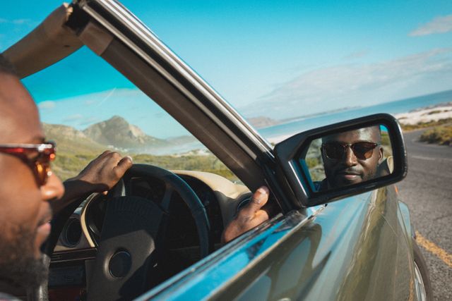 African american man smiling wearing sun glasses looking in wing mirror in convertible car. summer holiday road trip on a country highway by the coast.