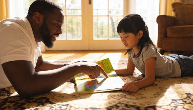Sunlit father reading picture book with daughter on cozy living room rug, storytime bonding