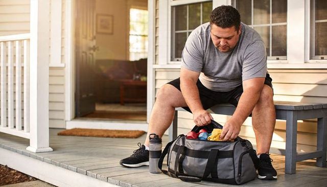Man packing gym bag on sunlit porch grabbing dumbbells and shaker bottle before workout