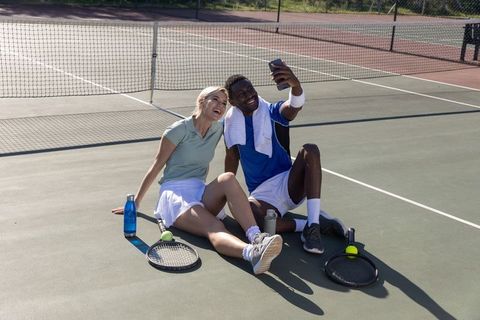 Smiling black man and white woman taking selfie while resting on outdoor tennis court