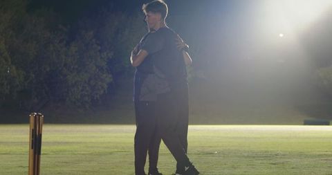 Night cricket teammates hugging under floodlights celebrating victory on grass field