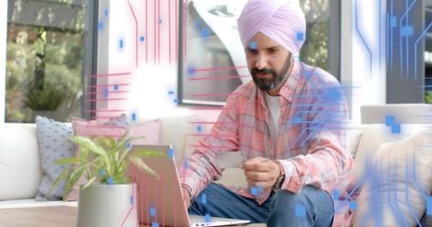 Man in turban holding card and using laptop on sofa for fintech online shopping
