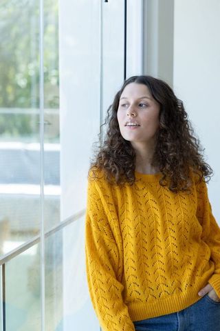 Young woman daydreaming by large glass window wearing mustard knit sweater and jeans